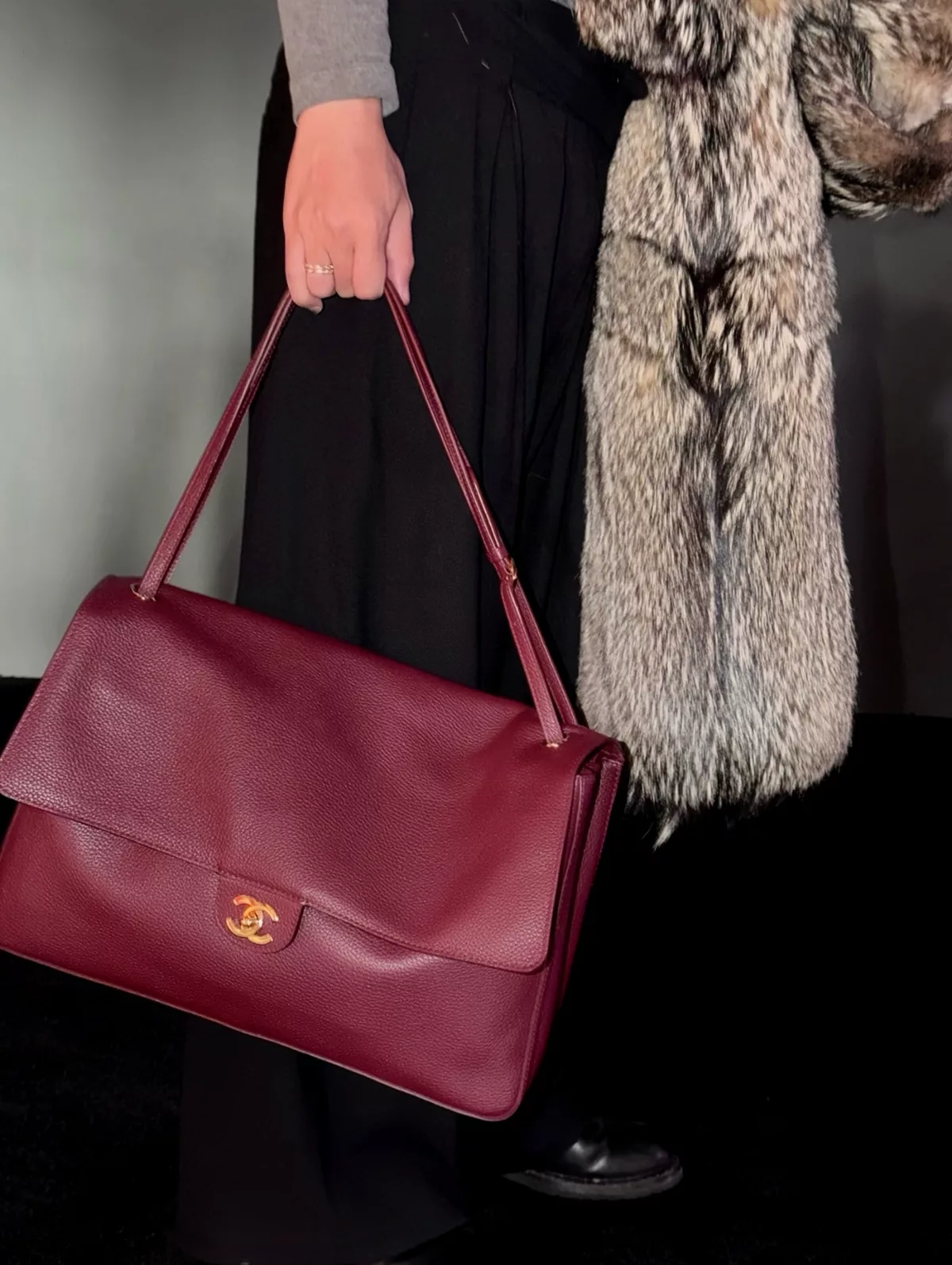 Burgundy flap shoulder bag against a grey sleeve and black trousers